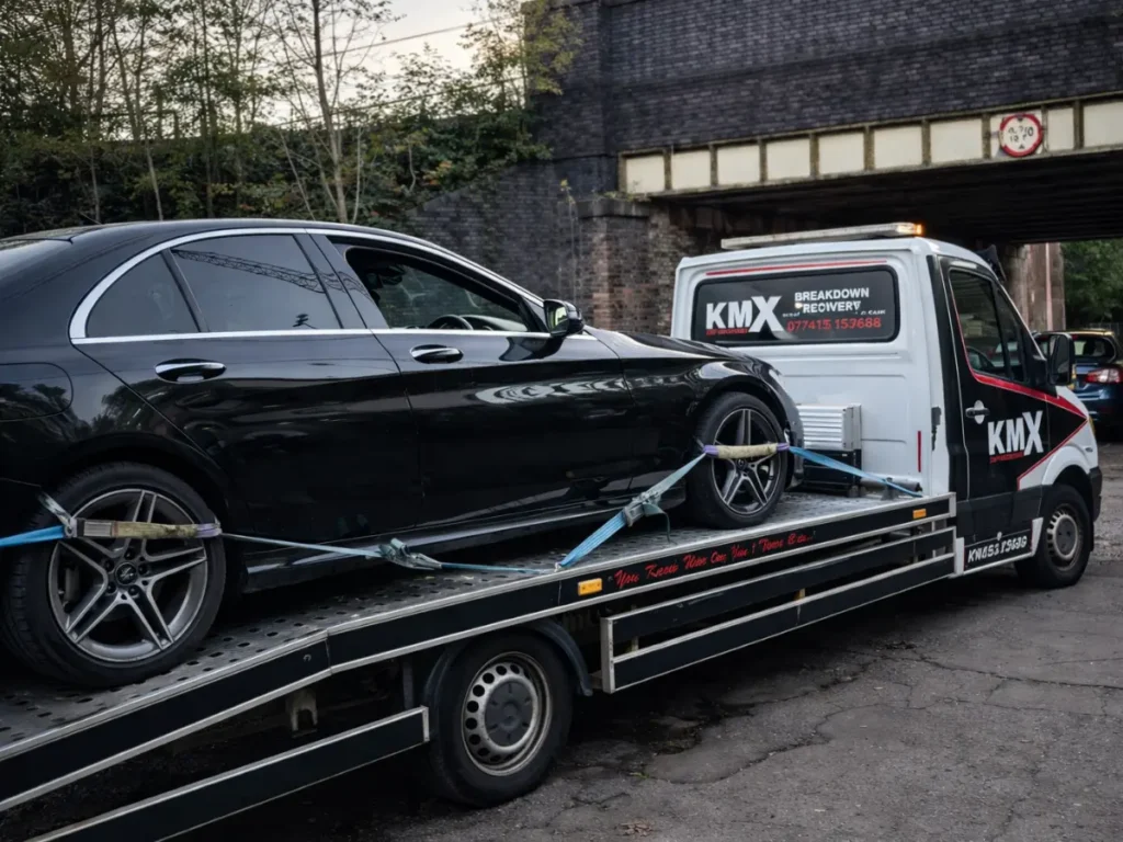 KMX recovery truck transporting a car on a flatbed under a bridge, demonstrating professional vehicle recovery service and safe car transport after breakdown