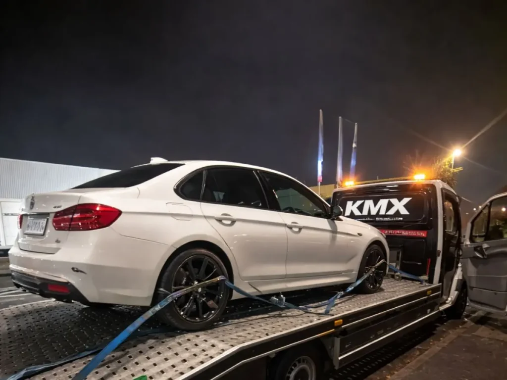 KMX recovery truck carrying a white car on a flatbed at night with secure straps and flashing lights